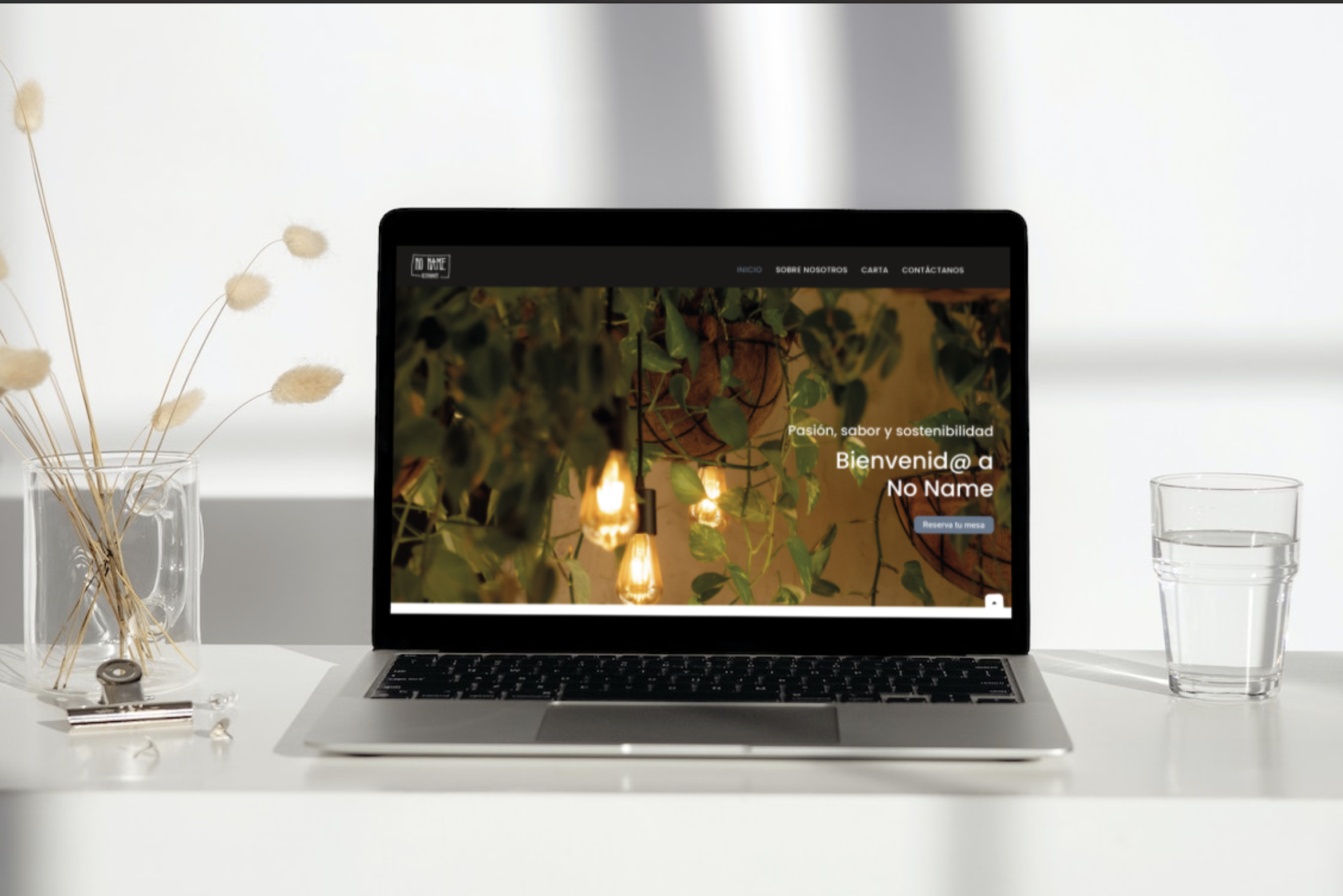 Mockup de la web del restaurante No Name sobre un escritorio limpio con un jarrón y un vaso de agua